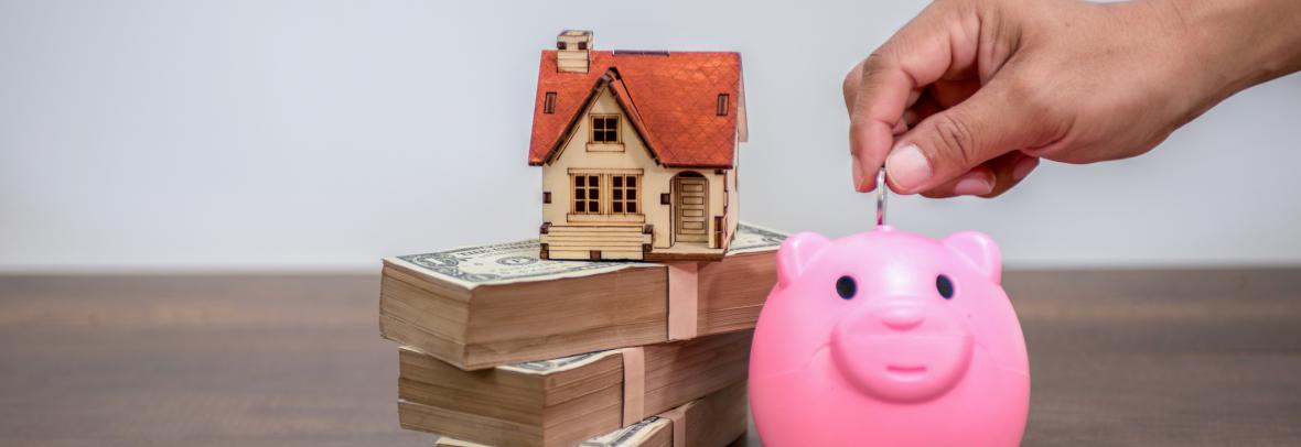 Toy home on top of money beside a piggy bank
