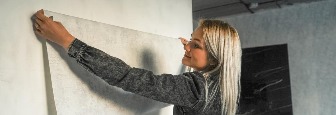 A young woman in a gray jumpsuit makes a modern renovation in a loft style. A woman tries on wallpaper against the wall.