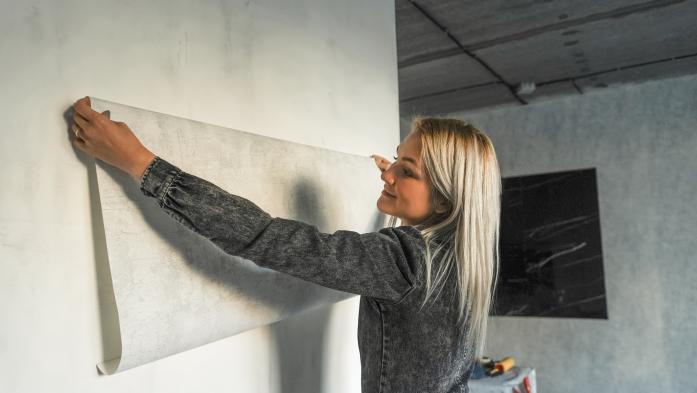 A young woman in a gray jumpsuit makes a modern renovation in a loft style. A woman tries on wallpaper against the wall.